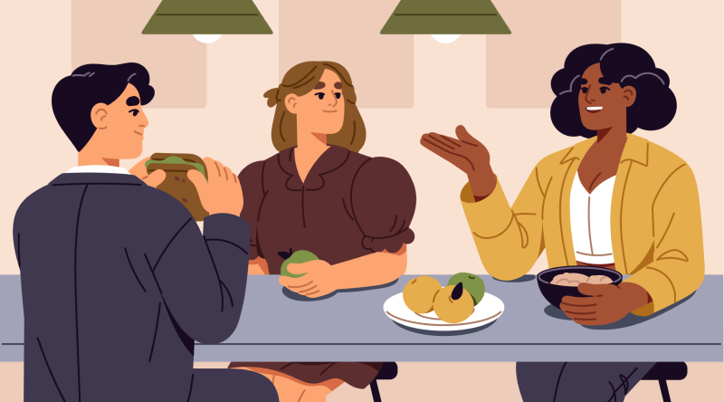 Two women and a man sat around a table talking. The man is wearing a suit and eating a sandwich. One woman has one hand on a bowl of food and the other held out gesturing as she speaks. The other woman is looking at the first woman and holding a green fruit. In the centre of the table is some fruit on a plate.